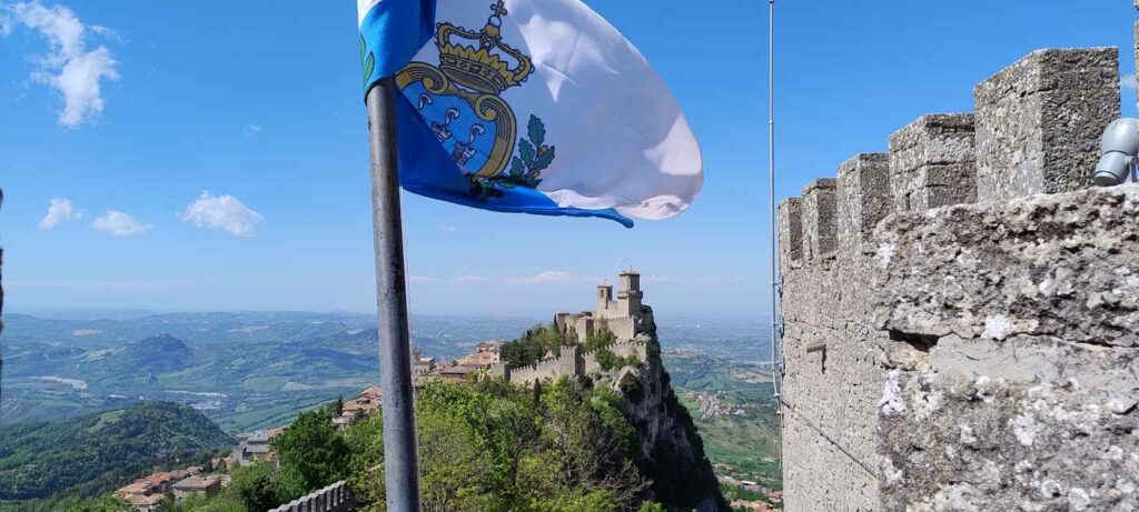 Blick auf den Torro Primo in San Marino. Der Turm steht auf der Spitze eines Berges, dahinter geht es mehrere hundert Meter nach unten. Im Vordergrund weht die Flagge von San Marino und die Burgmauer des Torro Secundo ist zu sehen.